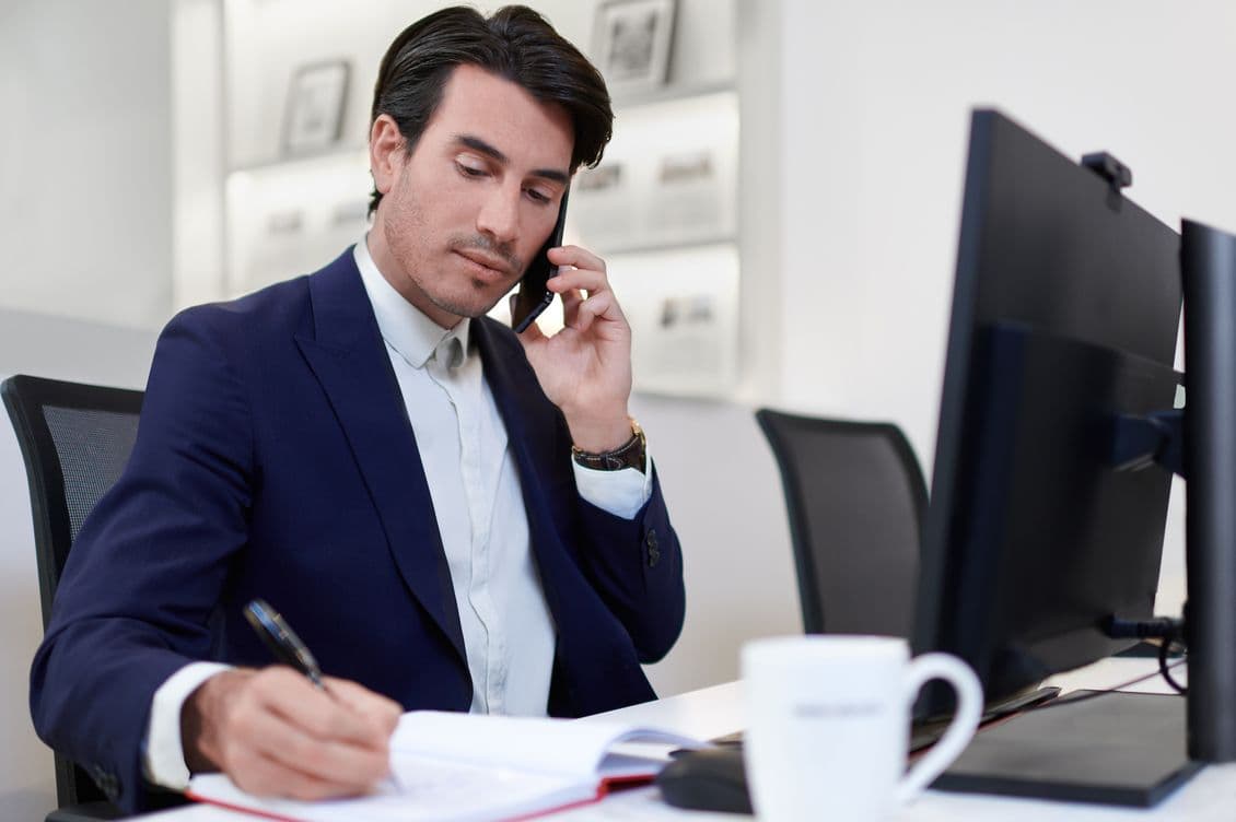Man in a suit talks on the phone while writing in a notebook at a desk with a computer and coffee mug in an office.