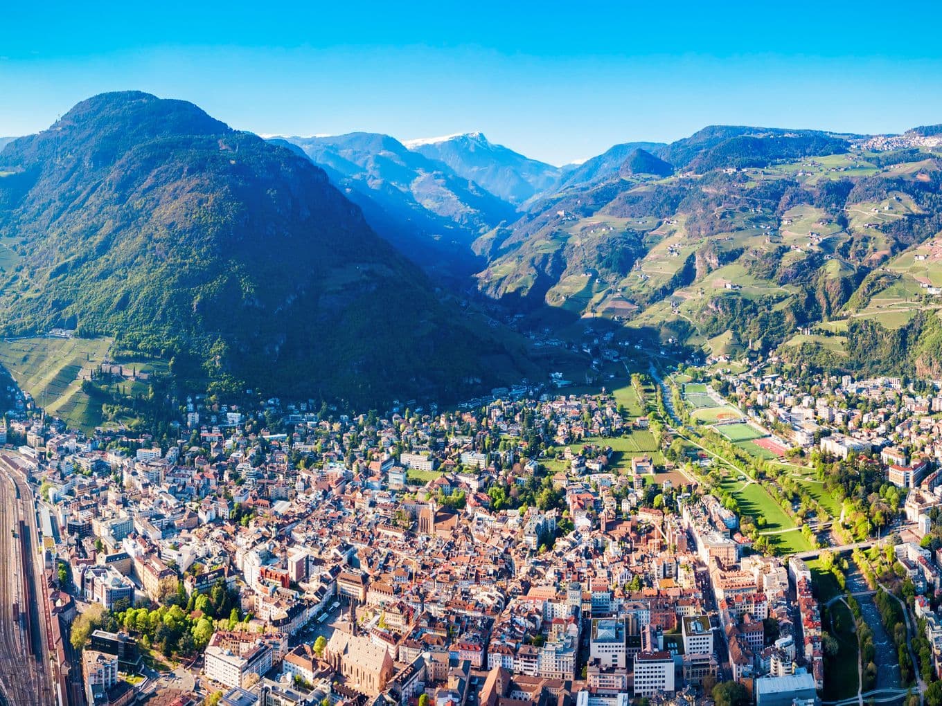 Luftaufnahme einer Stadt, eingebettet in ein Tal, umgeben von üppig grünen Bergen unter einem klaren blauen Himmel.