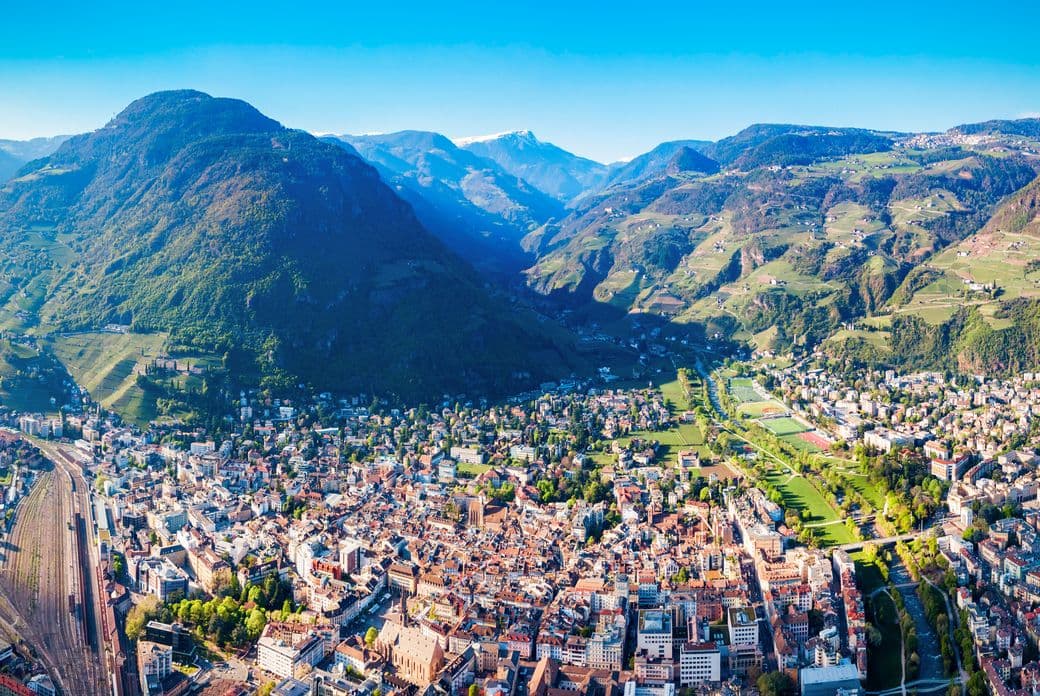 Aerial view of a city nestled in a mountainous valley, showcasing dense urban landscape, surrounding green hills, and clear blue skies.