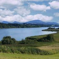 Panoramablick über den Wallersee mit grünen Ufern, Wiesen im Vordergrund und Bergkette im Hintergrund unter blauem Himmel mit weißen Wolken.