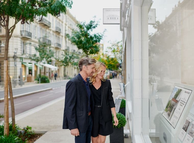 A couple in formal attire looks at real estate listings in a shop window on a tree-lined city street.