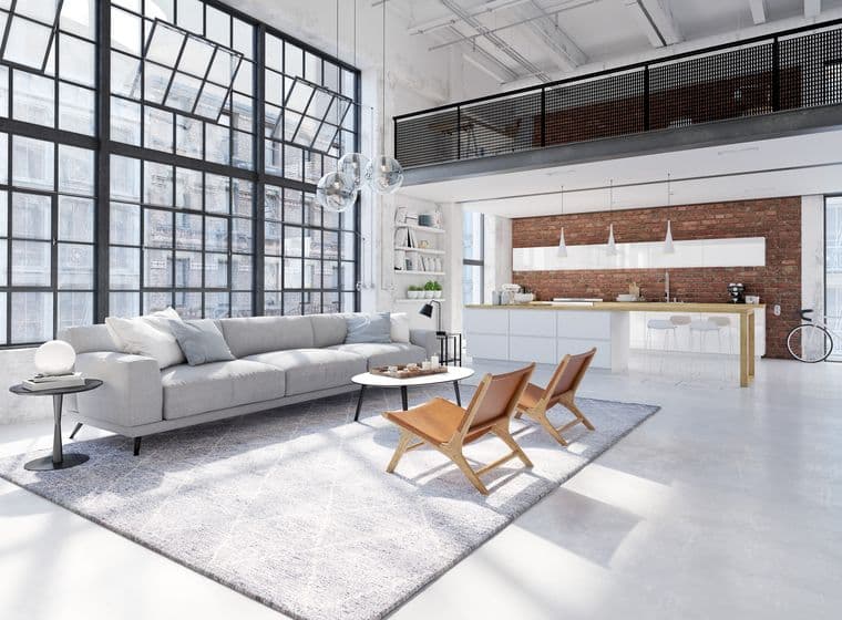 Bright, modern loft apartment with gray sofa, leather chairs, and large windows. A kitchen with brick wall is in the background.