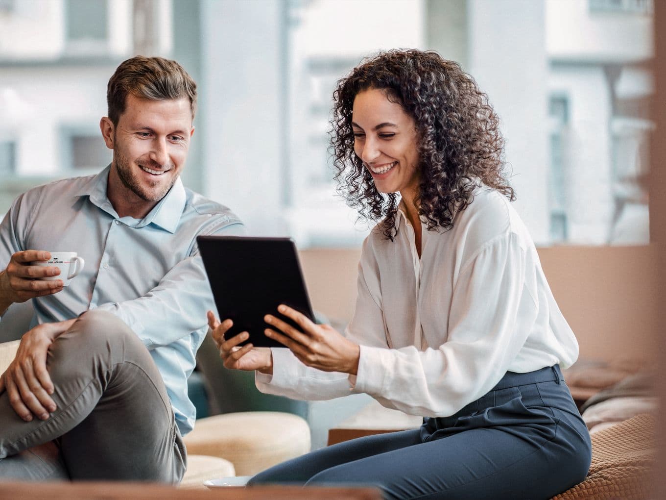 A man and woman sit together, smiling while looking at a tablet. The man holds a cup, and they appear to be in a casual, modern setting.