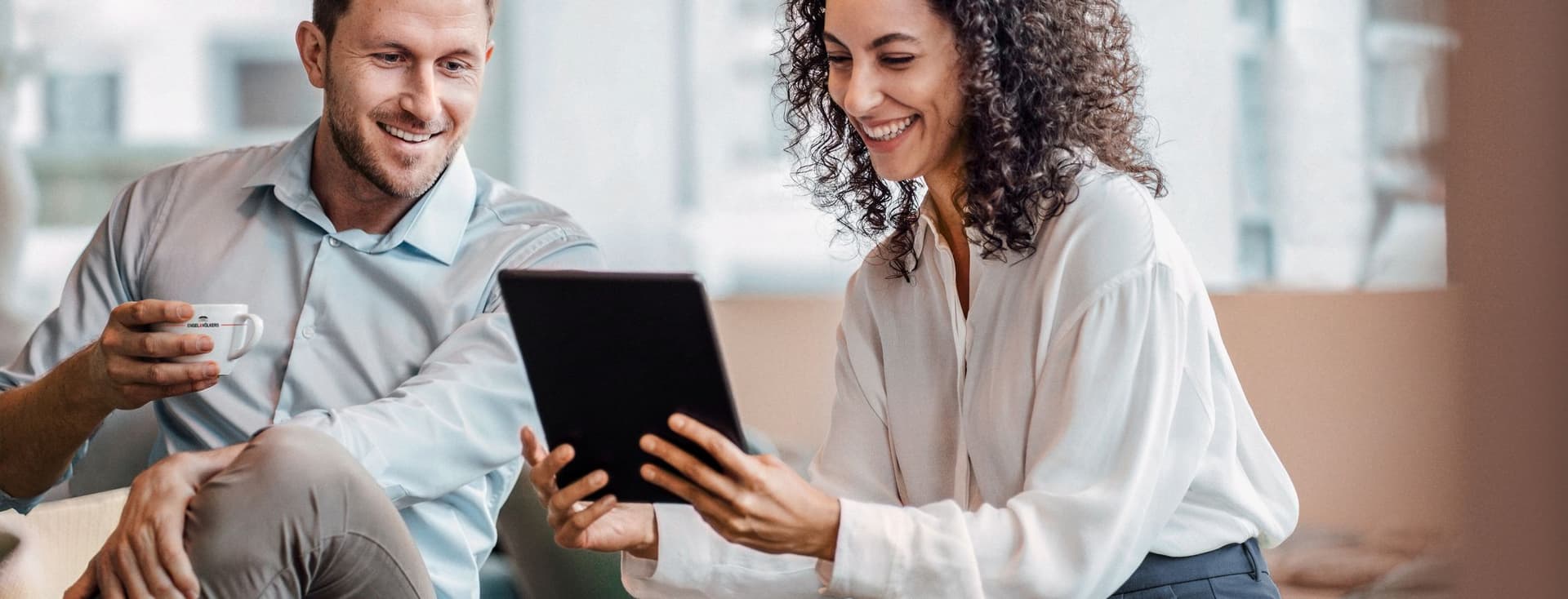 Ein Mann und eine Frau sitzen zusammen, lächeln und schauen auf ein Tablet. Der Mann hält eine Tasse in der Hand, und sie scheinen sich in einer lässigen, modernen Umgebung zu befinden.