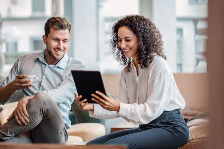 Ein Mann und eine Frau sitzen zusammen, lächeln und schauen auf ein Tablet. Der Mann hält eine Tasse in der Hand, und sie scheinen sich in einer lässigen, modernen Umgebung zu befinden.