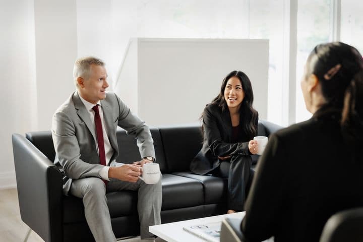 Two business people in suits sit on a black couch, holding mugs, and talking in a bright, modern office.