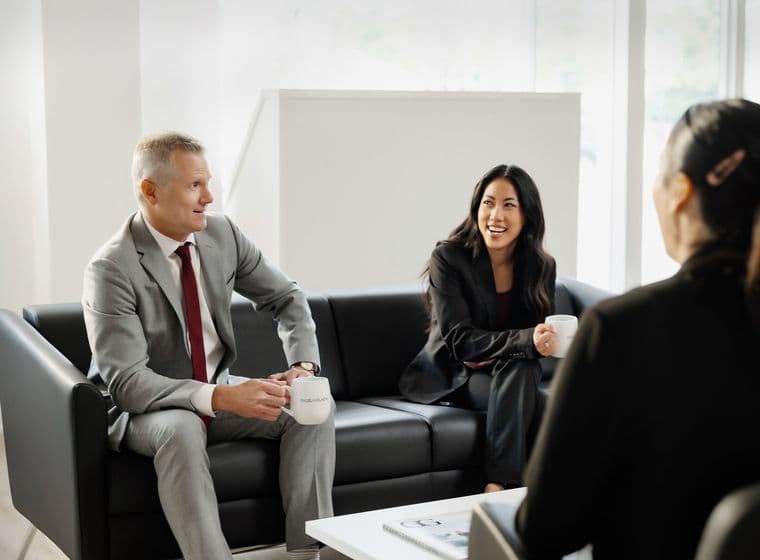 Two business people in suits sit on a black couch, holding mugs, and talking in a bright, modern office.