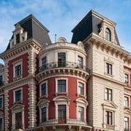 Historisches Mehrfamilienhaus im europäischen Stil mit verschnörkelten architektonischen Details, roter und cremefarbener Fassade, Bogenfenstern und schwarzen, schmiedeeisernen Balkonen unter blauem Himmel.