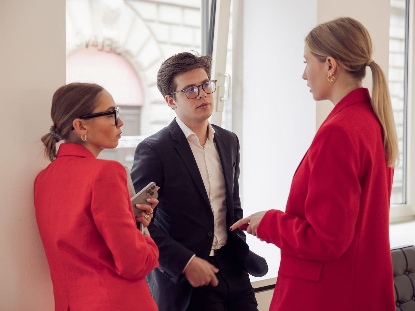 Three professionals in formal attire, two women in red jackets and a man in a black suit, engaged in conversation by a window.