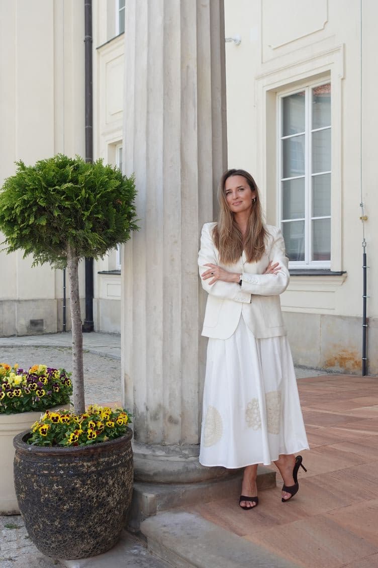 Woman in a cream blazer and white skirt stands with arms crossed beside a tall column outside a pale building, near potted flowers.