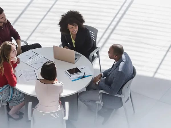 Six diverse professionals sit around a round table in a bright, modern office, collaborating with laptops, tablets, and documents.