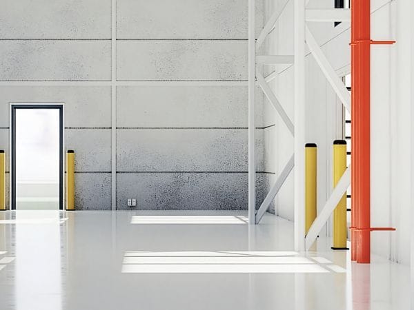 Interior of a spacious, empty warehouse with white walls, a concrete floor, and large doors, featuring yellow and red safety bollards.