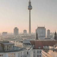 Panoramic Berlin skyline at sunrise with the TV tower and cathedral dome rising above historic rooftops under a pastel sky.
