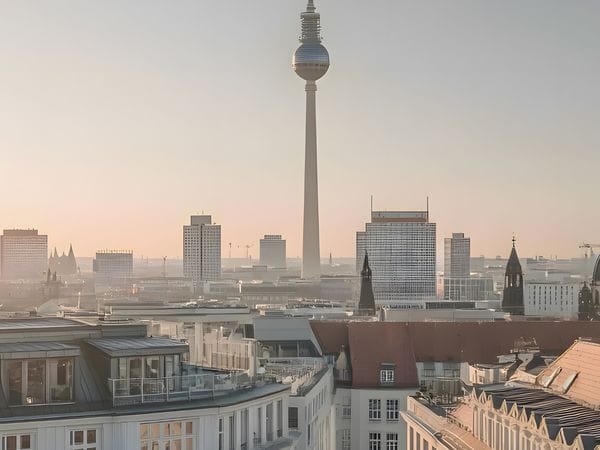 Panoramic Berlin skyline at sunrise with the TV tower and cathedral dome rising above historic rooftops under a pastel sky.