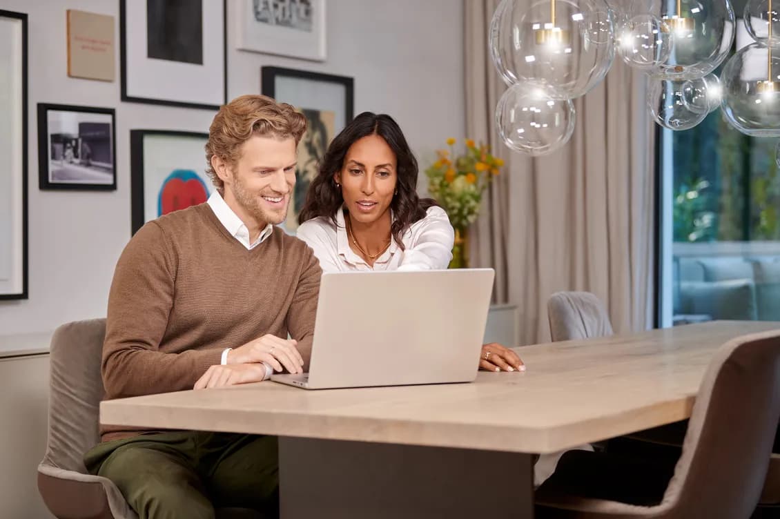 A man and woman sit at a table, looking at a laptop together, with framed art on the wall and a vase of flowers in the background.