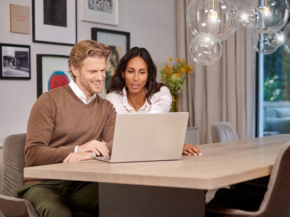 A man and woman sit at a table, looking at a laptop together, with framed art on the wall and a vase of flowers in the background.