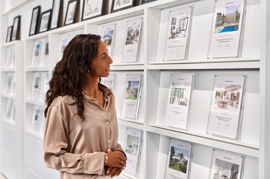 A woman in a beige blouse looks at real estate listings displayed on a white wall in a modern office setting.