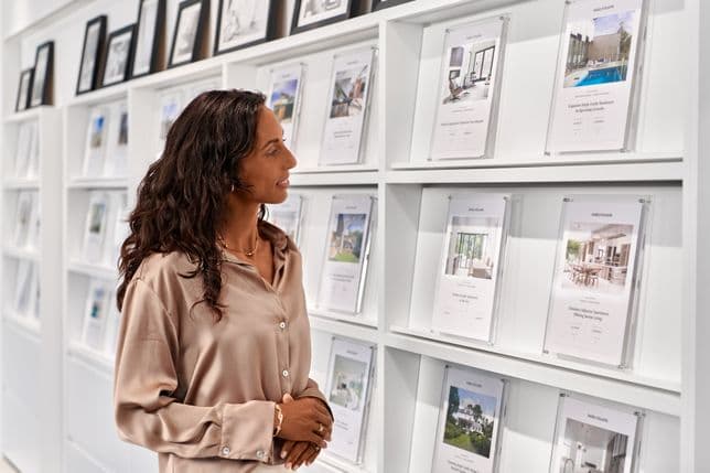 A woman in a beige blouse looks at real estate listings displayed on a white wall in a modern office setting.