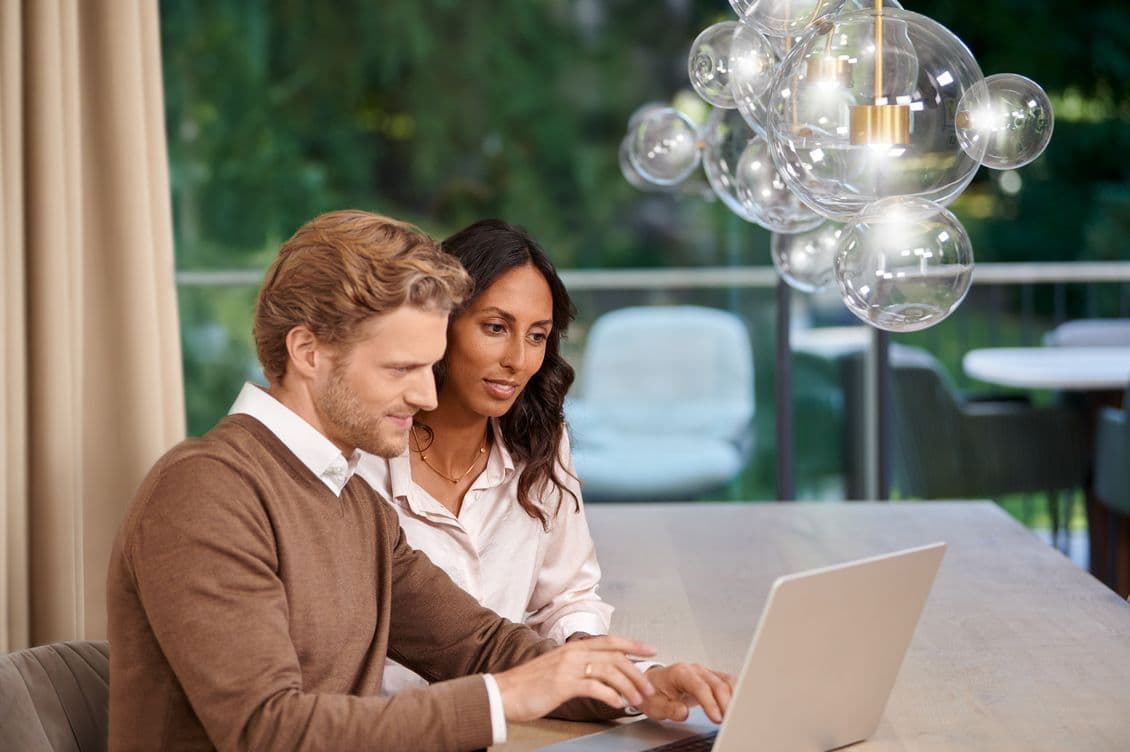 A man and woman sit at a table, focused on a laptop. They are in a modern room with a glass bubble chandelier and a view of greenery outside.