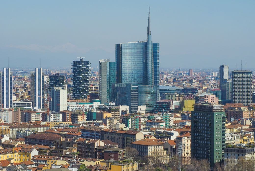 Aerial view of a modern cityscape featuring tall skyscrapers, residential buildings with red roofs, and clear blue sky overhead.