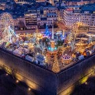 Aerial view of a vibrant, illuminated Christmas market at night, featuring a Ferris wheel, carousel, and decorated trees in a city setting.
