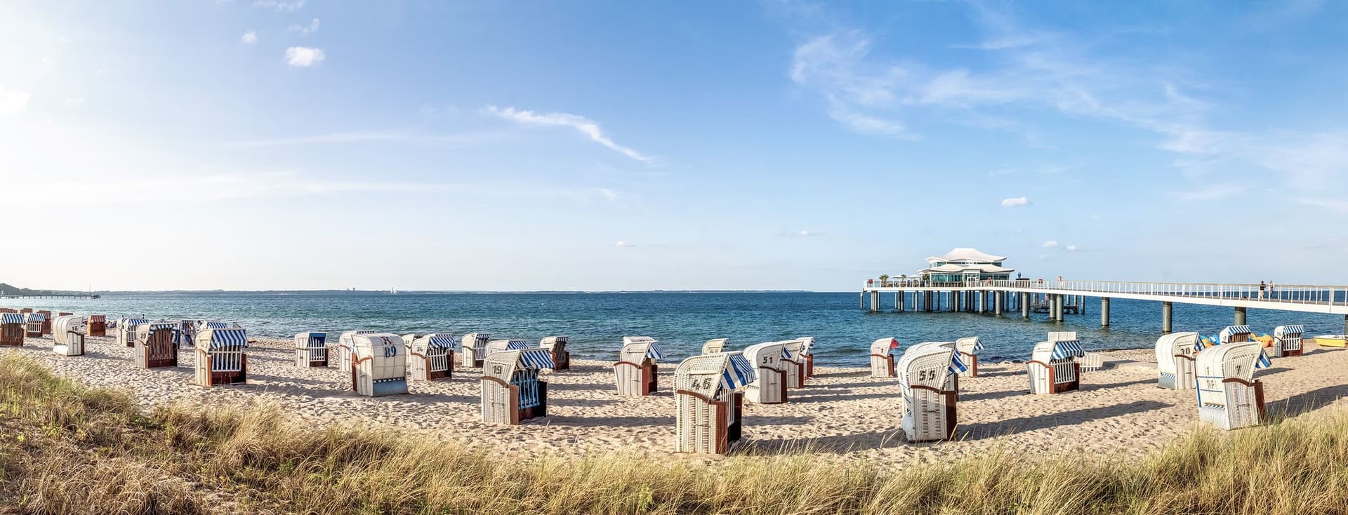 Beach scene with many striped beach chairs on sand, blue sea, and a pier with a white building at the end under a blue sky.