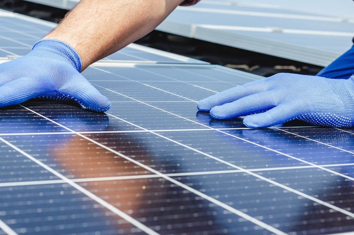 Close-up of hands wearing blue protective gloves working on solar panels, showing professional installation or maintenance of photovoltaic modules with reflective surface.