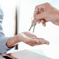 Close-up of hands during property key handover, one hand wearing a blue business shirt sleeve passing keys to another hand wearing a gray sleeve, symbolizing successful real estate transaction completion