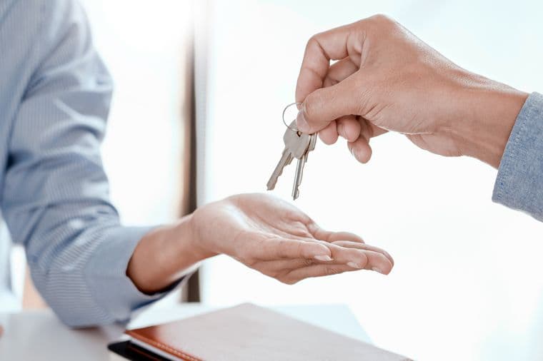 Close-up of hands during property key handover, one hand wearing a blue business shirt sleeve passing keys to another hand wearing a gray sleeve, symbolizing successful real estate transaction completion