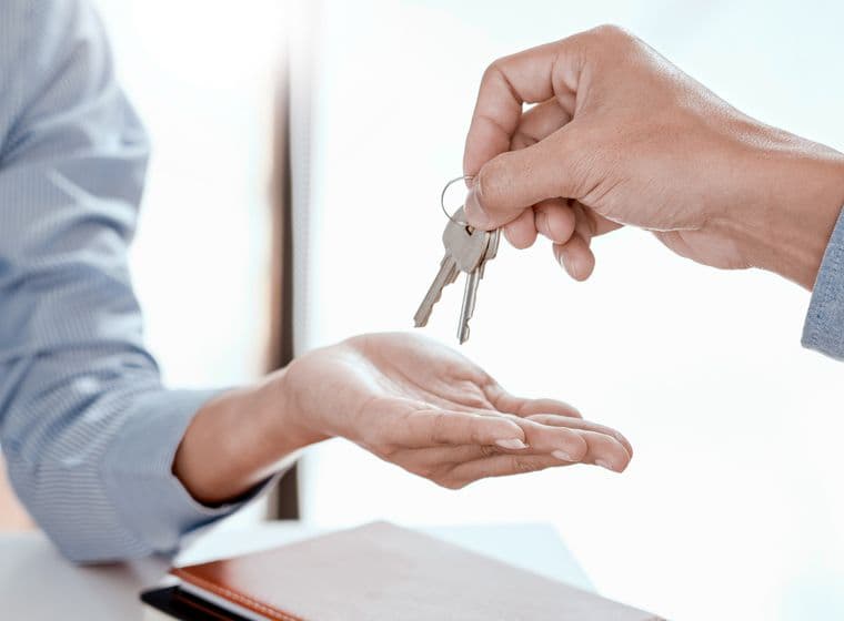 Close-up of hands during property key handover, one hand wearing a blue business shirt sleeve passing keys to another hand wearing a gray sleeve, symbolizing successful real estate transaction completion