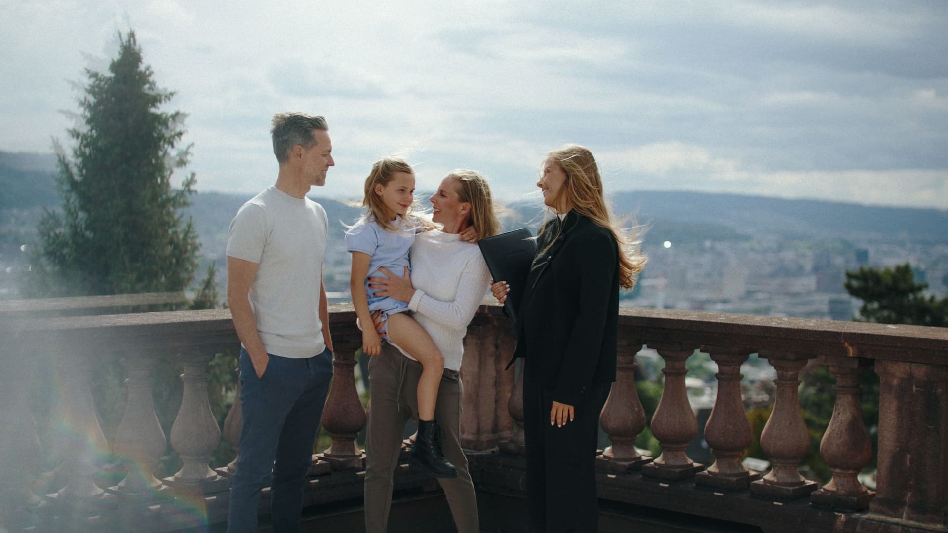 Family of four on a rooftop balcony overlooking a city and hills, smiling; man in a light shirt, woman holding a young girl, and another woman with a folder.