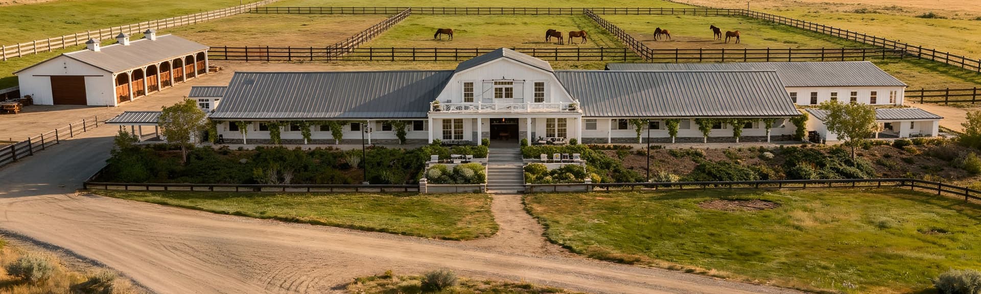 Aerial view of a large ranch with a white farmhouse, fenced fields, horses grazing, and mountains in the background.