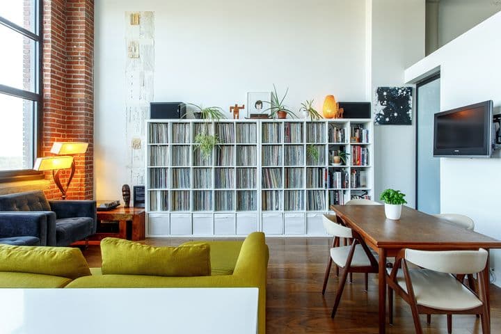 The picture shows a living room with a green sofa and wooden table/chairs with light-coloured seats and backrests. There is a large white bookcase against the wall. There is a window on the left.