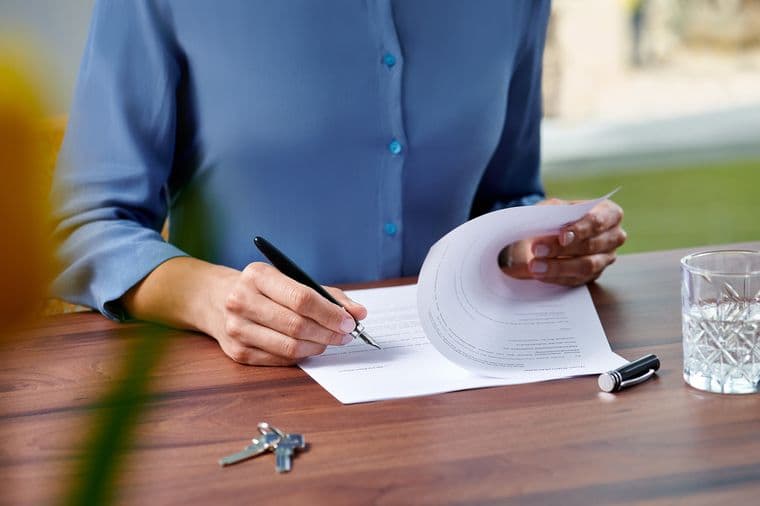 Person in blue blouse reviewing and signing papers at wooden desk with keys and crystal glass visible, photographed in a warm, natural setting