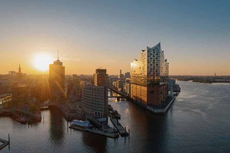 The image shows the impressive architecture with its undulating glass roof, reminiscent of water waves or sails. The lower third of the building consists of a robust red brick base, reminiscent of the former quay warehouse building. The Elbe river can be seen in the foreground.