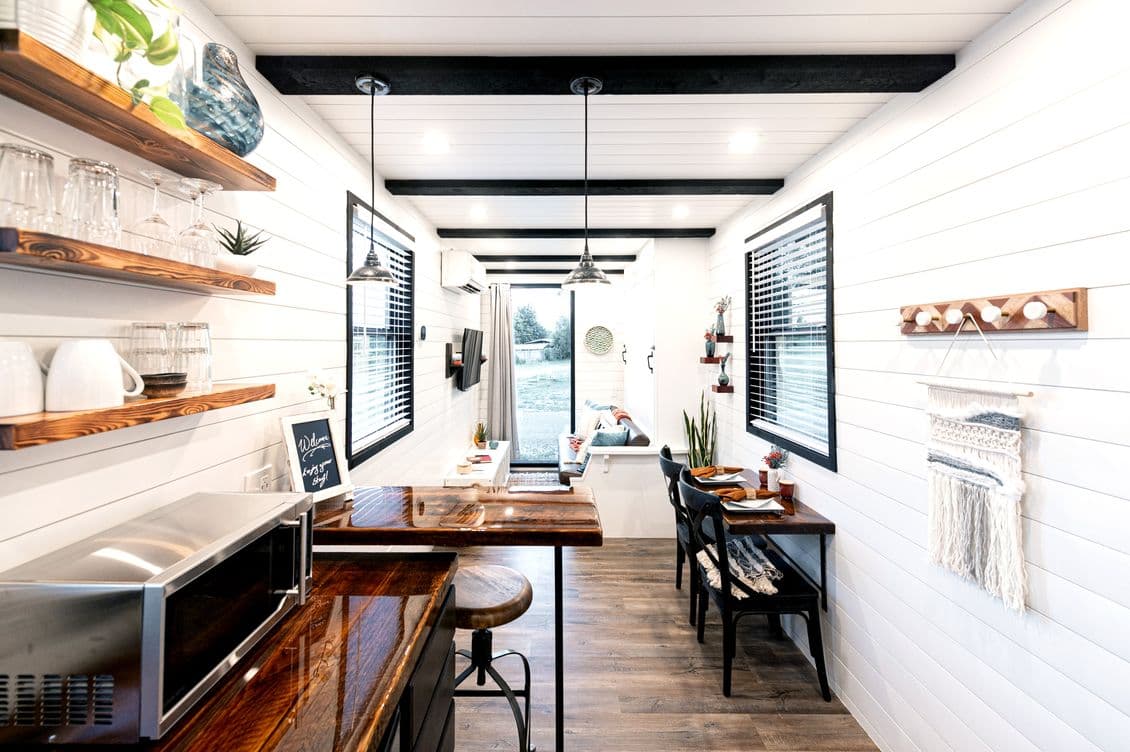 A beautifully designed tiny house featuring a modern kitchen with wooden countertops, open shelving and industrial pendant lighting. The open-plan layout leads to a cosy living area with large windows, creating a bright and inviting atmosphere. AI-generated image