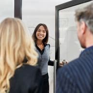 A woman smiling and welcoming two people at a doorway, one with blonde hair, the other with gray hair, in a professional setting.