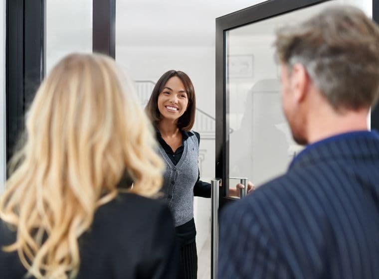 A woman smiling and welcoming two people at a doorway, one with blonde hair, the other with gray hair, in a professional setting.