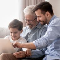 Ein kleiner Junge, sein Vater und sein Großvater sitzen zusammen auf einem grauen Sofa und schauen lächelnd auf ein Tablet, mit dem sie interagieren. Sie sind leger gekleidet: Der Großvater trägt eine Brille und ein graues Hemd, der Vater ein hellblaues Hemd und der Junge ein weißes T-Shirt.