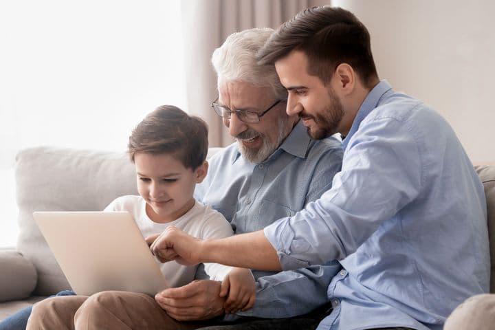 Ein kleiner Junge, sein Vater und sein Großvater sitzen zusammen auf einer grauen Couch und lächeln, während sie auf ein Tablet-Gerät schauen und mit diesem interagieren. Sie sind leger gekleidet, der Großvater trägt eine Brille und ein graues Hemd, der Vater ein hellblaues Hemd und der Junge ein weißes T-Shirt.