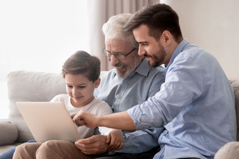 A young boy, his father, and grandfather sitting together on a grey couch, all smiling while looking at and interacting with a tablet device. They are dressed casually, with the grandfather wearing glasses and a grey shirt, the father in a light blue dress shirt, and the boy in a white t-shirt.