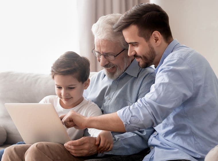 A young boy, his father, and grandfather sitting together on a grey couch, all smiling while looking at and interacting with a tablet device. They are dressed casually, with the grandfather wearing glasses and a grey shirt, the father in a light blue dress shirt, and the boy in a white t-shirt.