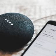 A black smart speaker next to a smartphone displaying a 'Welcome Home' screen with device setup interface. The smart speaker has illuminated indicator lights on top and a textured fabric covering.