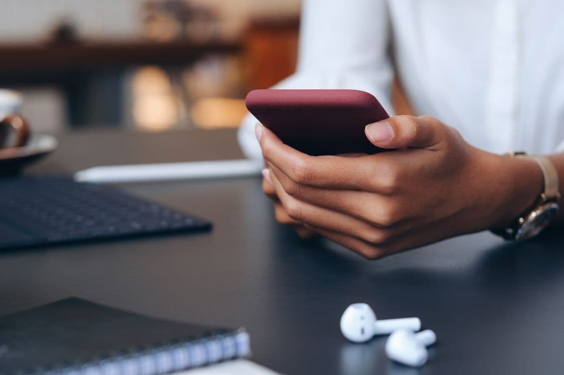 Close-up view of hands holding a smartphone with wireless earbuds and laptop keyboard visible on dark desk surface, depicting modern business communication setup