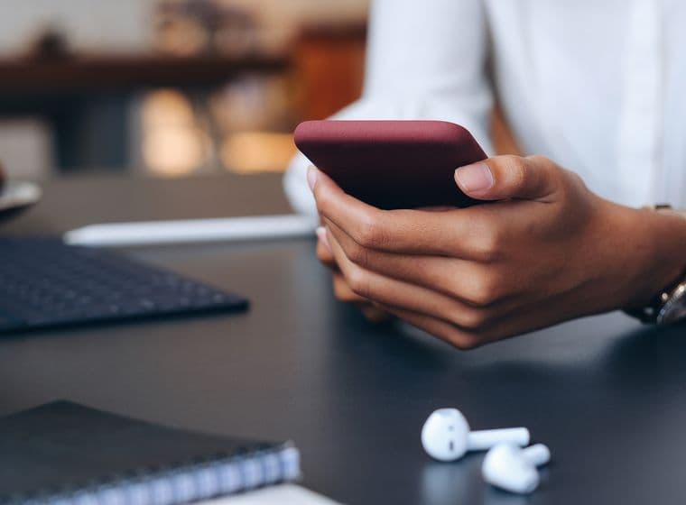 Close-up of hands holding a maroon phone, with white earbuds on a black desk. A notebook and keyboard are visible.