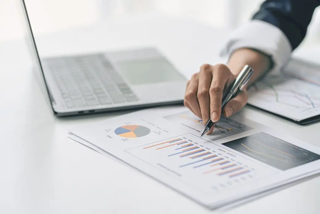 Hand holding pen over charts on a white desk with a laptop in the background.