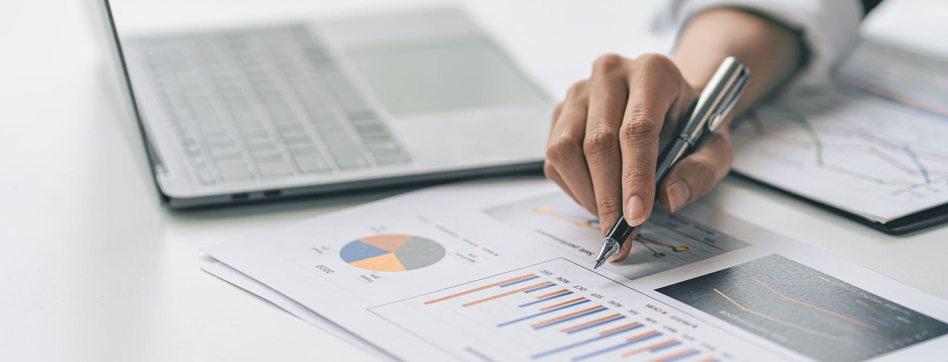 Close-up of a person's hand in business attire using a pen to analyze colorful charts and graphs on a financial report, with a laptop visible in the background on a white desk.