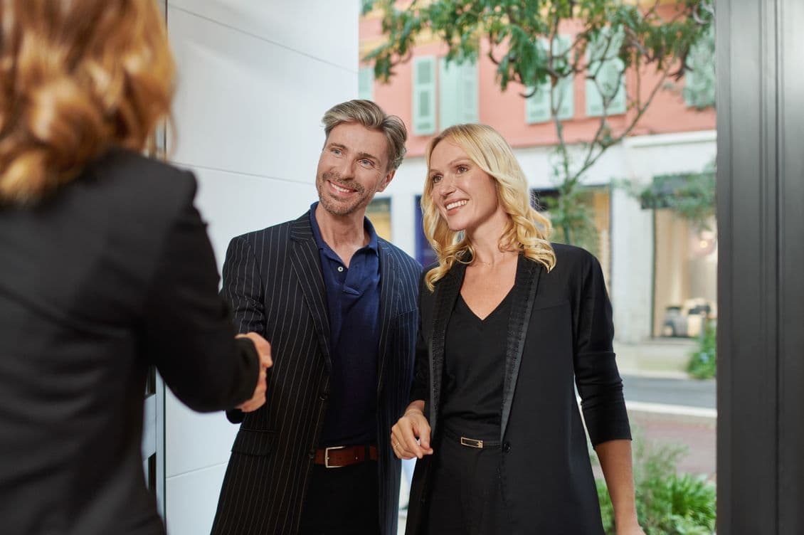 A smiling couple shakes hands with a person indoors, standing near a window with a view of a street and buildings outside.