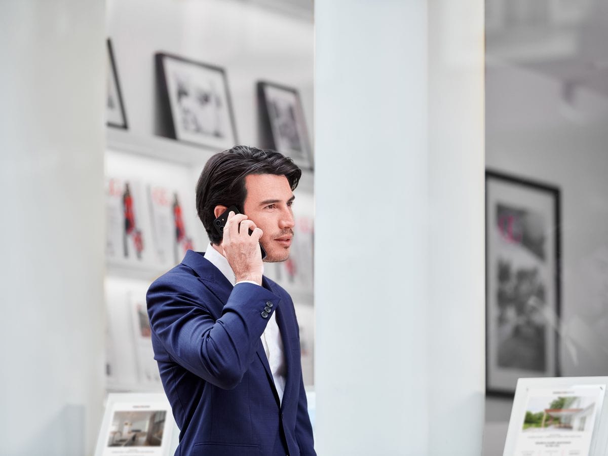 Man in a blue suit talking on a smartphone, standing in a modern office with framed pictures and brochures in the background.