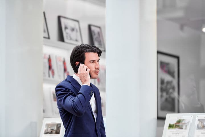 Man in a blue suit talking on a smartphone, standing in a modern office with framed pictures and brochures in the background.